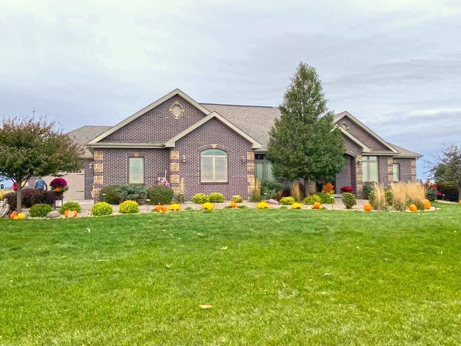 A single-story brick house with a brown roof and evergreen tree, surrounded by a green lawn and autumn landscaping.