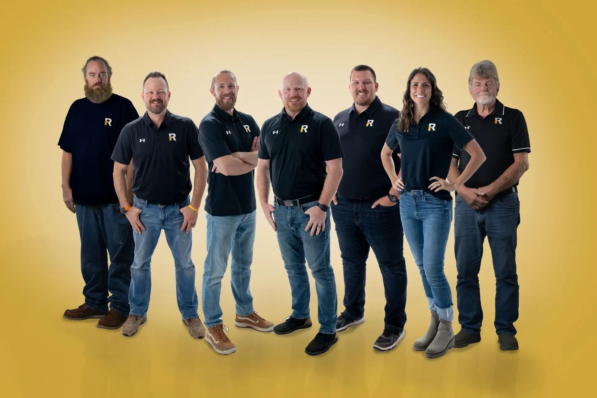 A group of seven people stand side-by-side in matching dark polo shirts and jeans against a solid gold background.