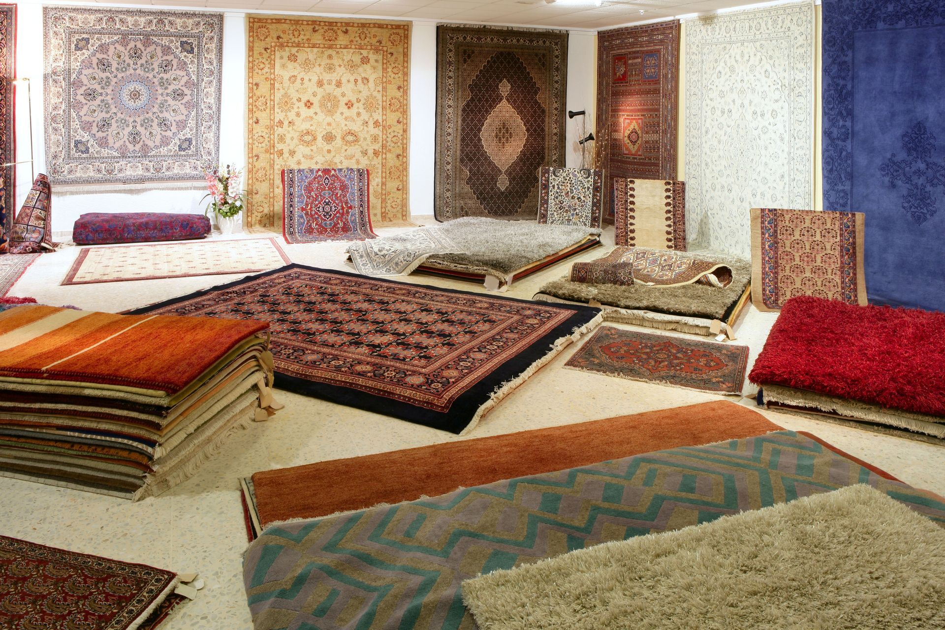 Carpets displayed in a store, various patterns and colors. Some stacked, some on the floor, and some on walls.