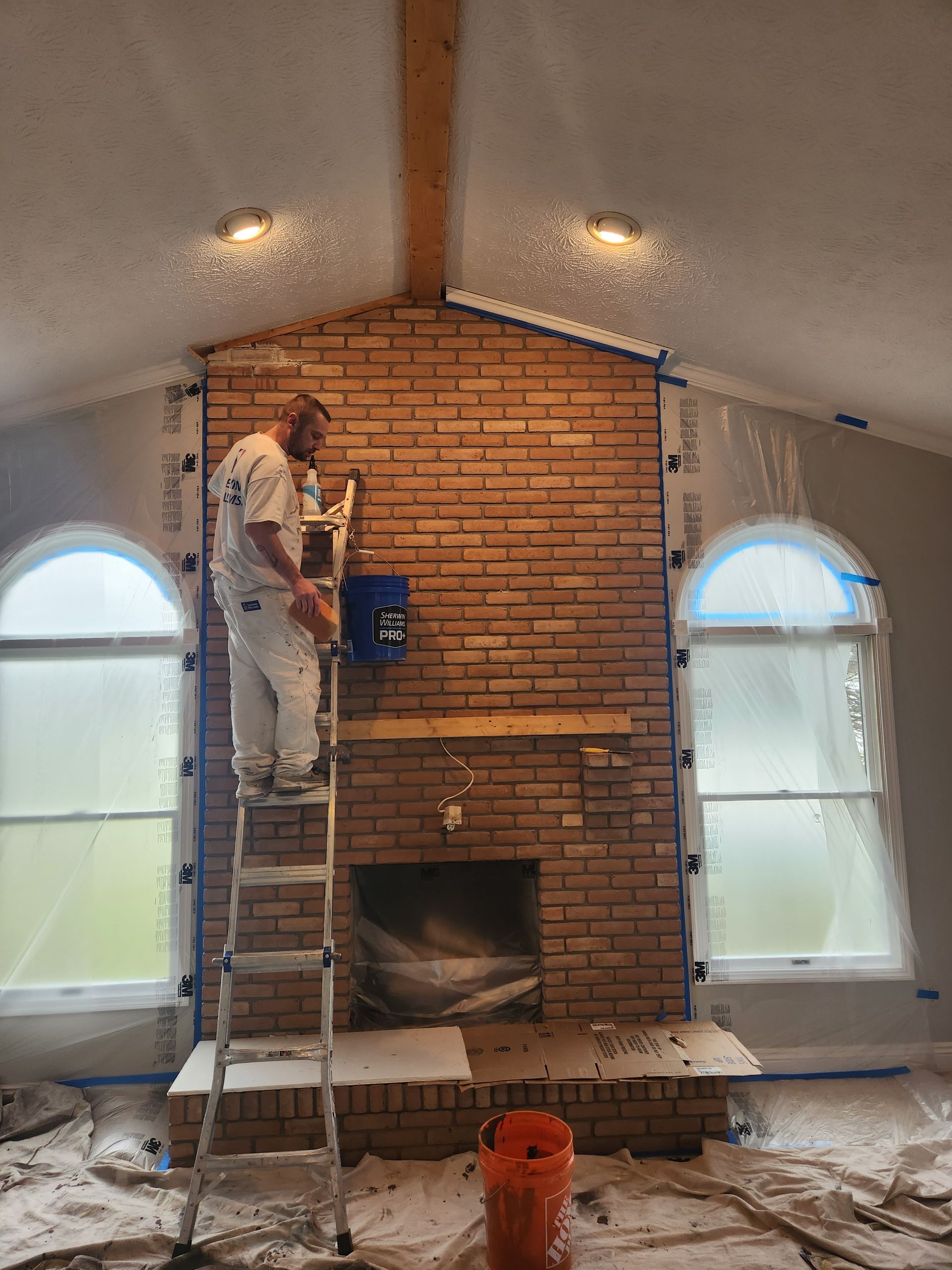 A man is standing on a ladder painting a brick fireplace in a living room.
