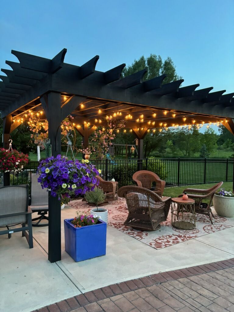 A patio with a pergola , chairs , tables and purple flowers.