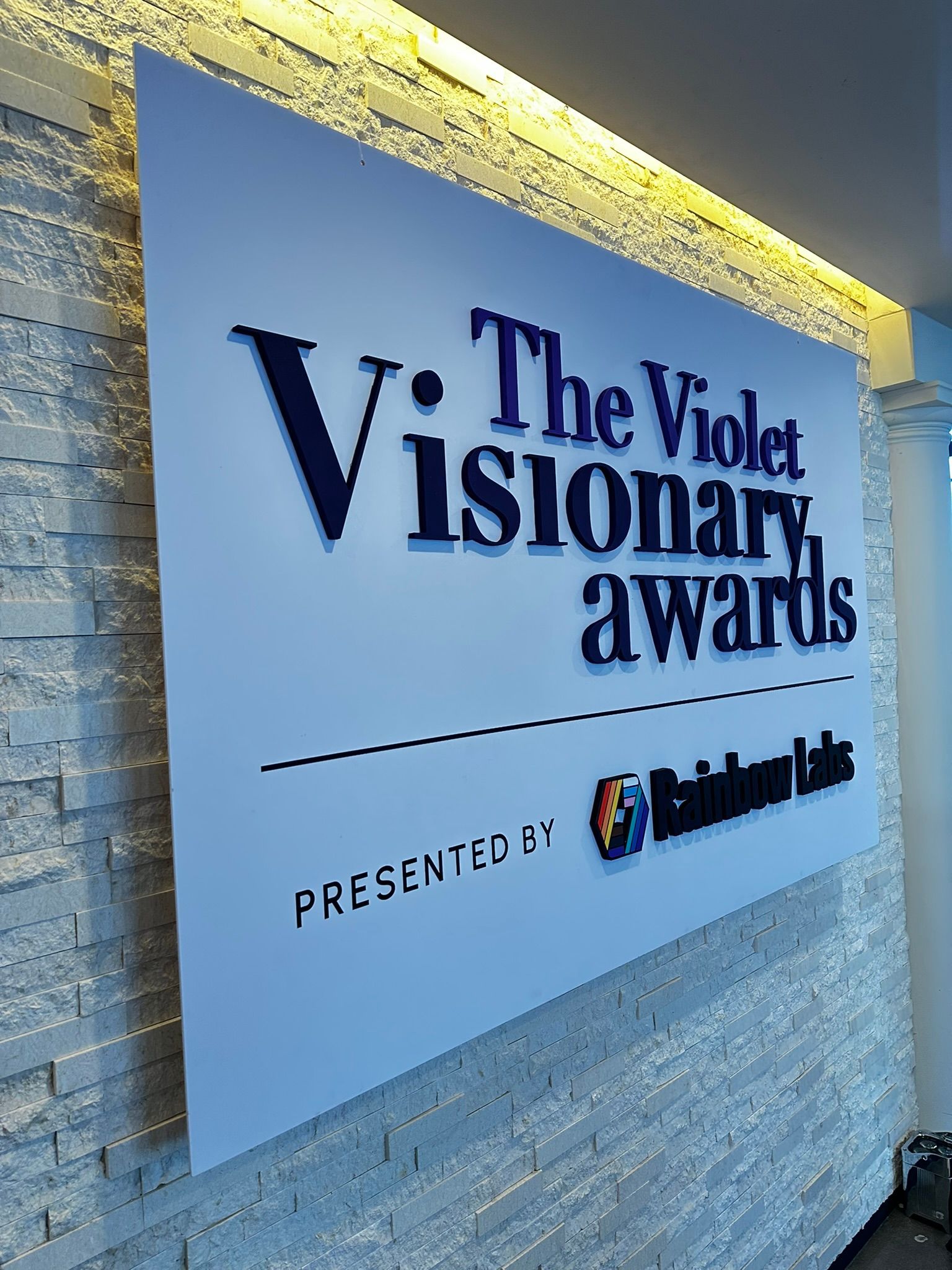 Sign for "The Violet Visionary Awards" presented by ReidHealth, against a light brick wall.