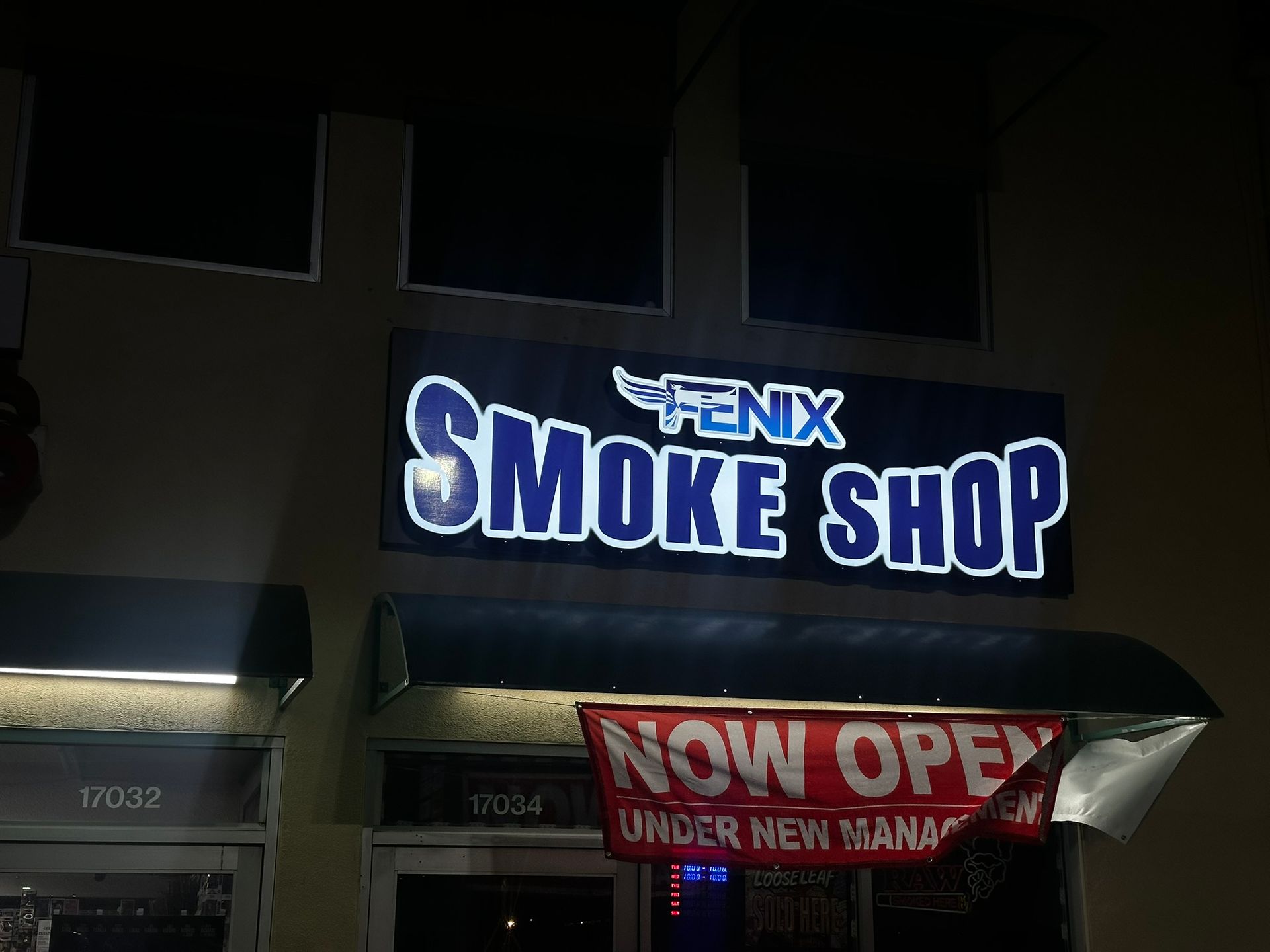 A lit sign for a smoke shop that says "FENIX Smoke Shop." A banner reads "NOW OPEN UNDER NEW MANAGEMENT."