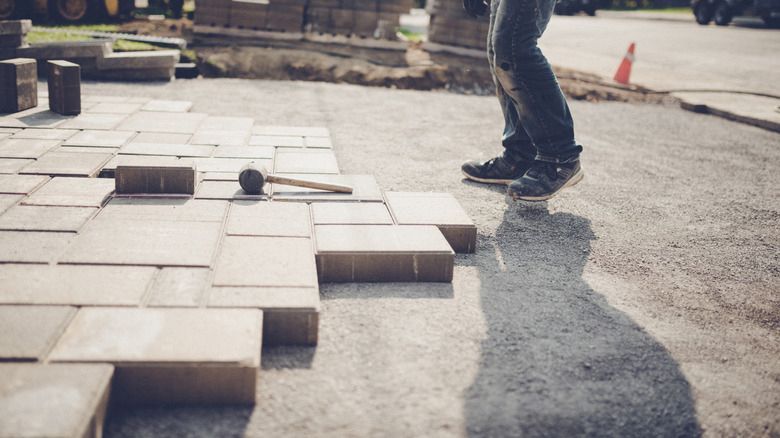 Person laying pavers on a paved surface. A mallet lies nearby.