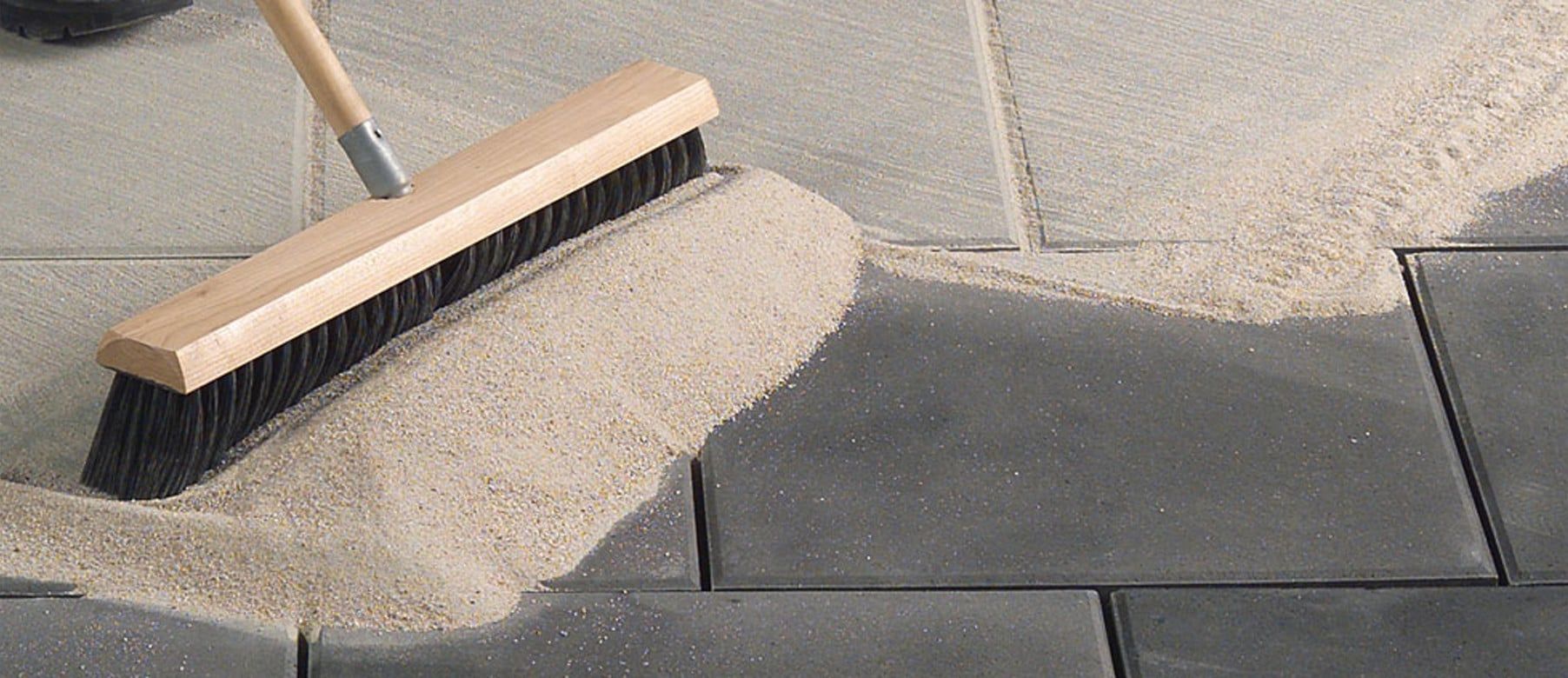 A person sweeping sand into the joints of paving stones with a large broom.