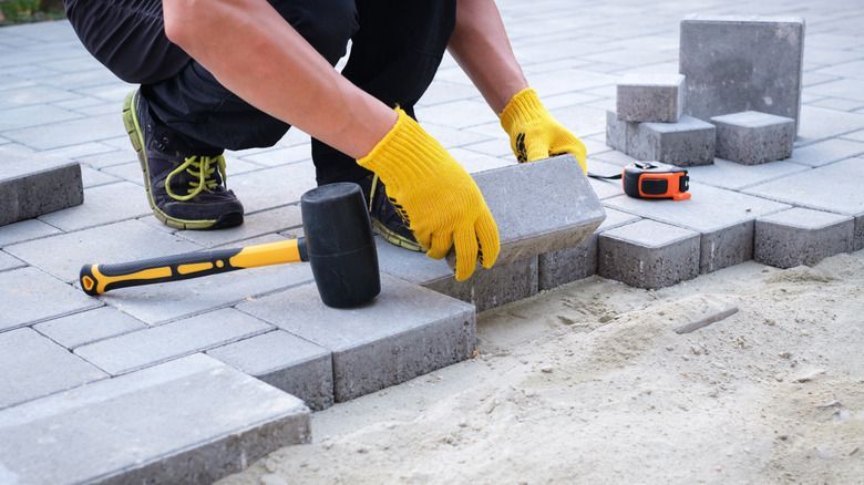 Person in yellow gloves installing paving stones with a mallet and measuring tape in an outdoor setting.