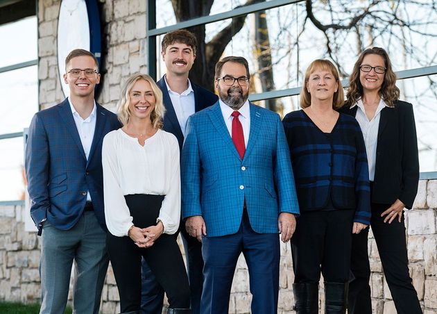 Six people standing outdoors in business attire in front of a building and trees.