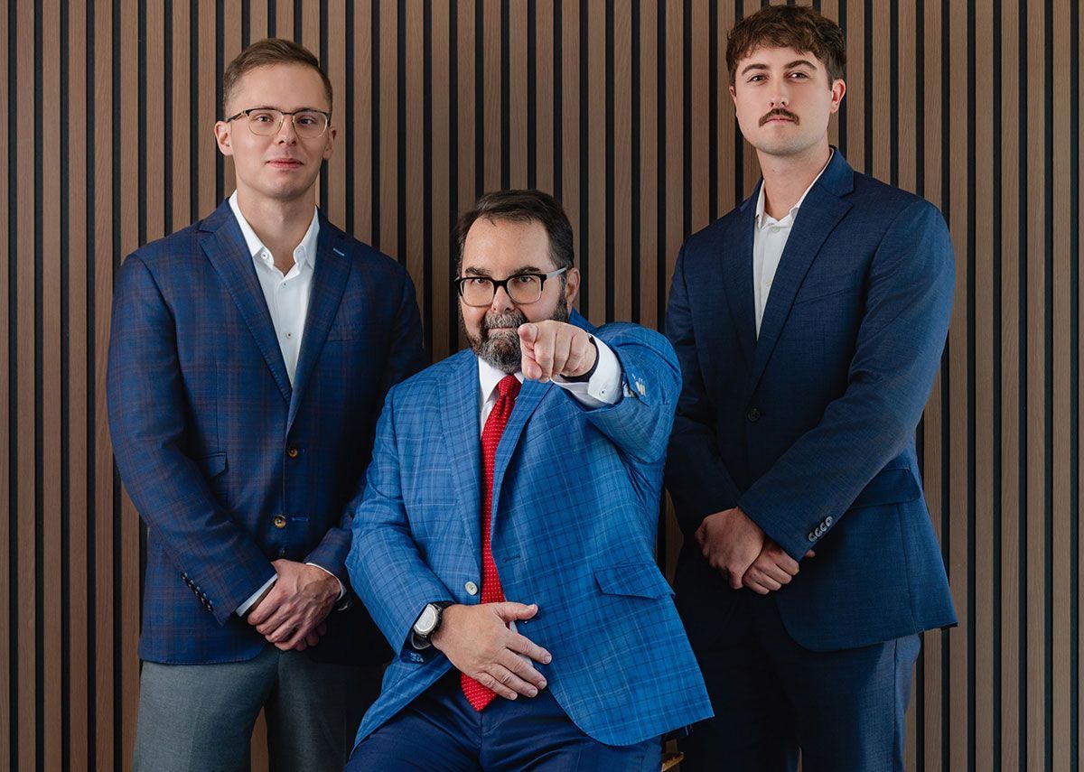 Three men in blue suits pose against a wood-paneled wall, with the center man pointing at the camera.