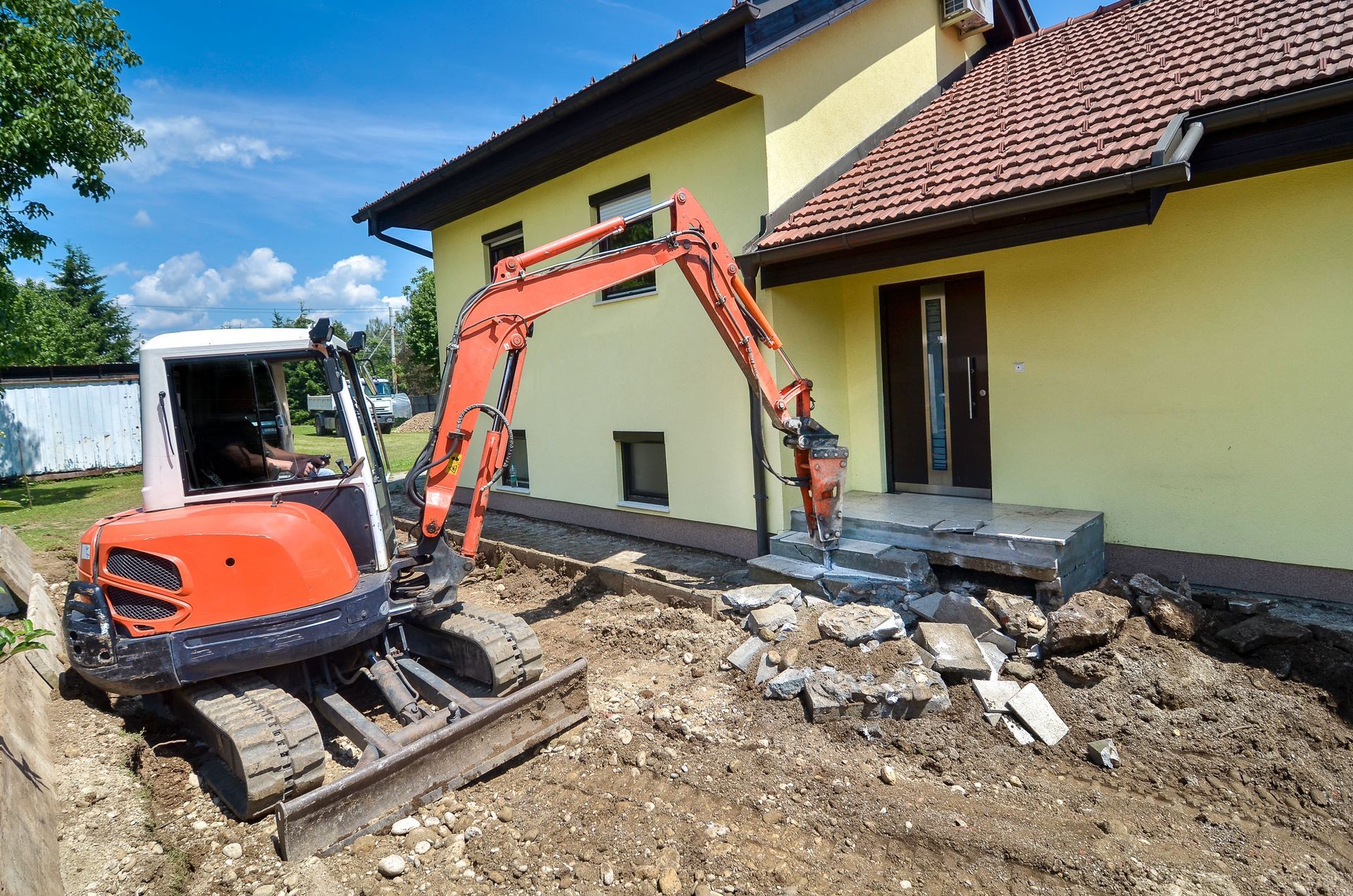 Mini excavator demolishing a concrete front step of a yellow house on a sunny day.