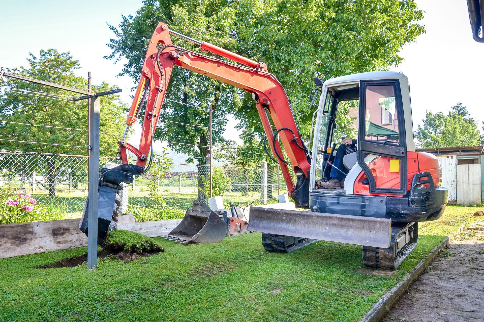 Orange excavator digging in a grassy yard, under a blue sky, near a clothesline and trees.