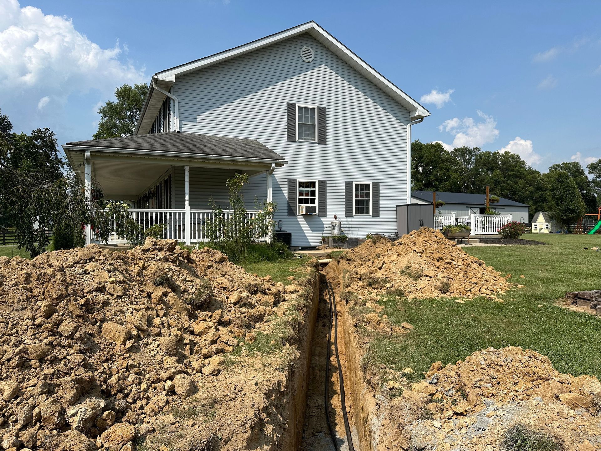 A trench dug in a yard in front of a two-story house with a porch.