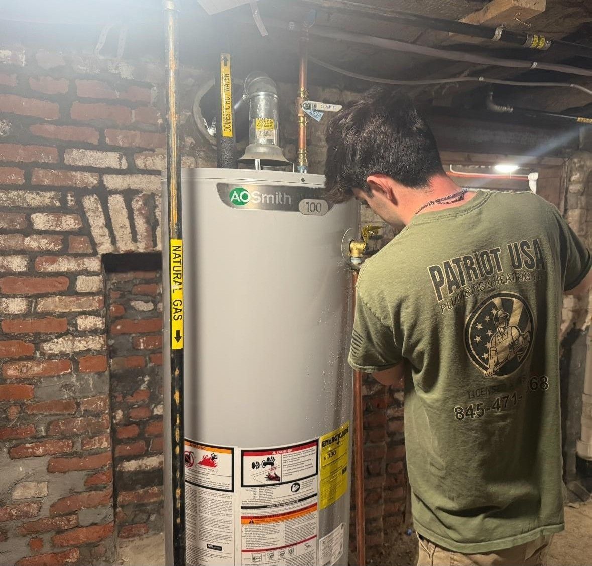 Man in green shirt inspecting a water heater in a basement next to a brick wall.