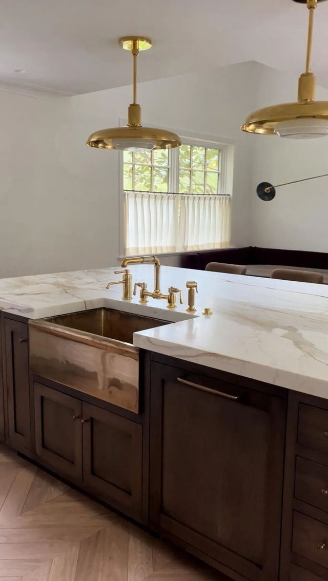Modern kitchen with dark wood island, white countertops, brass pendant lights, and a sink by a window.