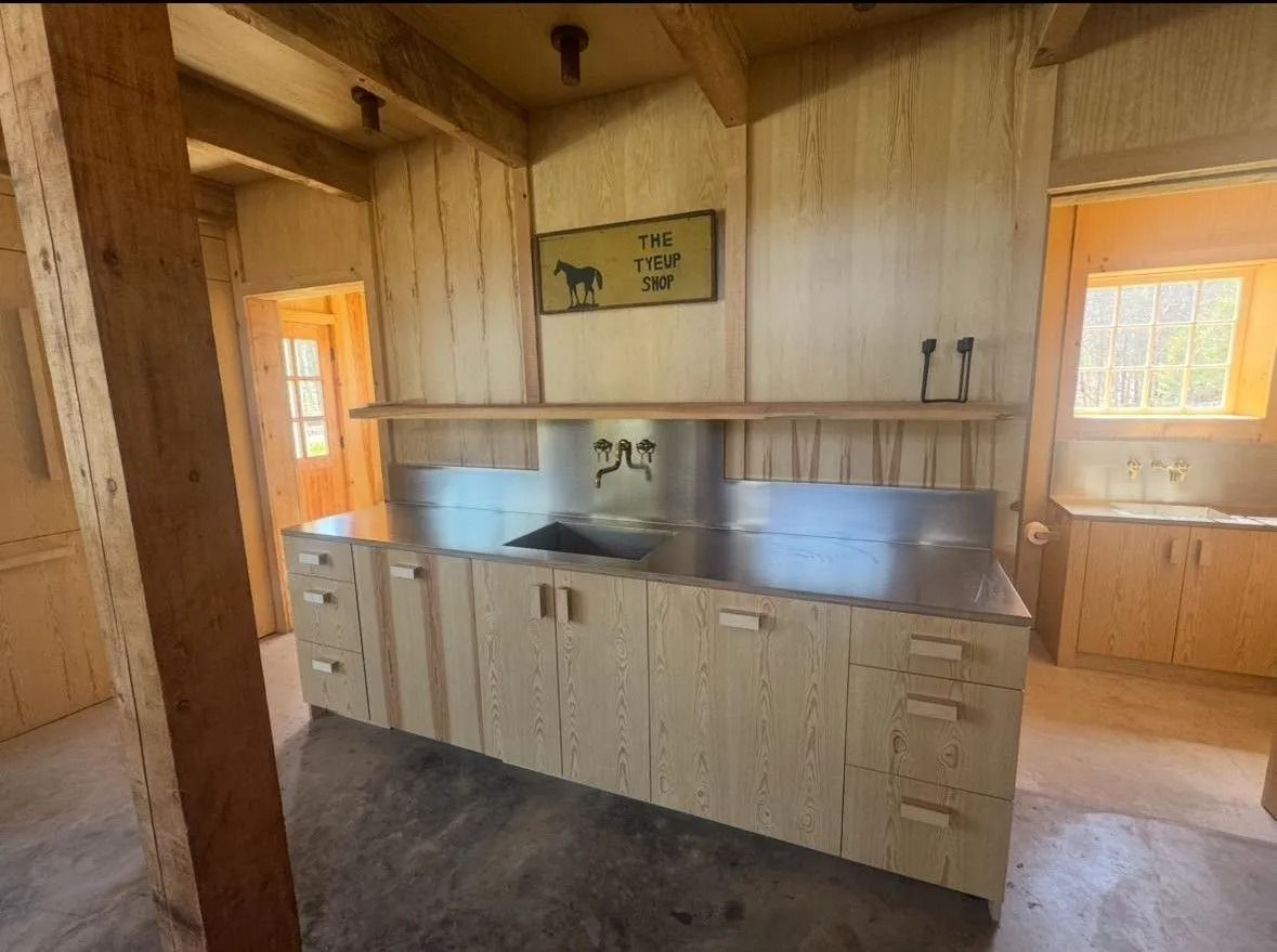 Rustic kitchen with wood-paneled walls, stainless counter, and light wood cabinets in an unfinished room