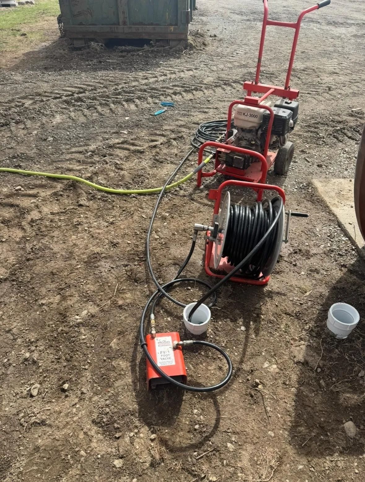Red drain-cleaning machine with hose reels on dirt beside a truck, with cups and a cable nearby