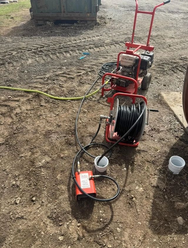 Red drain cleaning machine and hose reels on dirt ground with cables and buckets nearby.