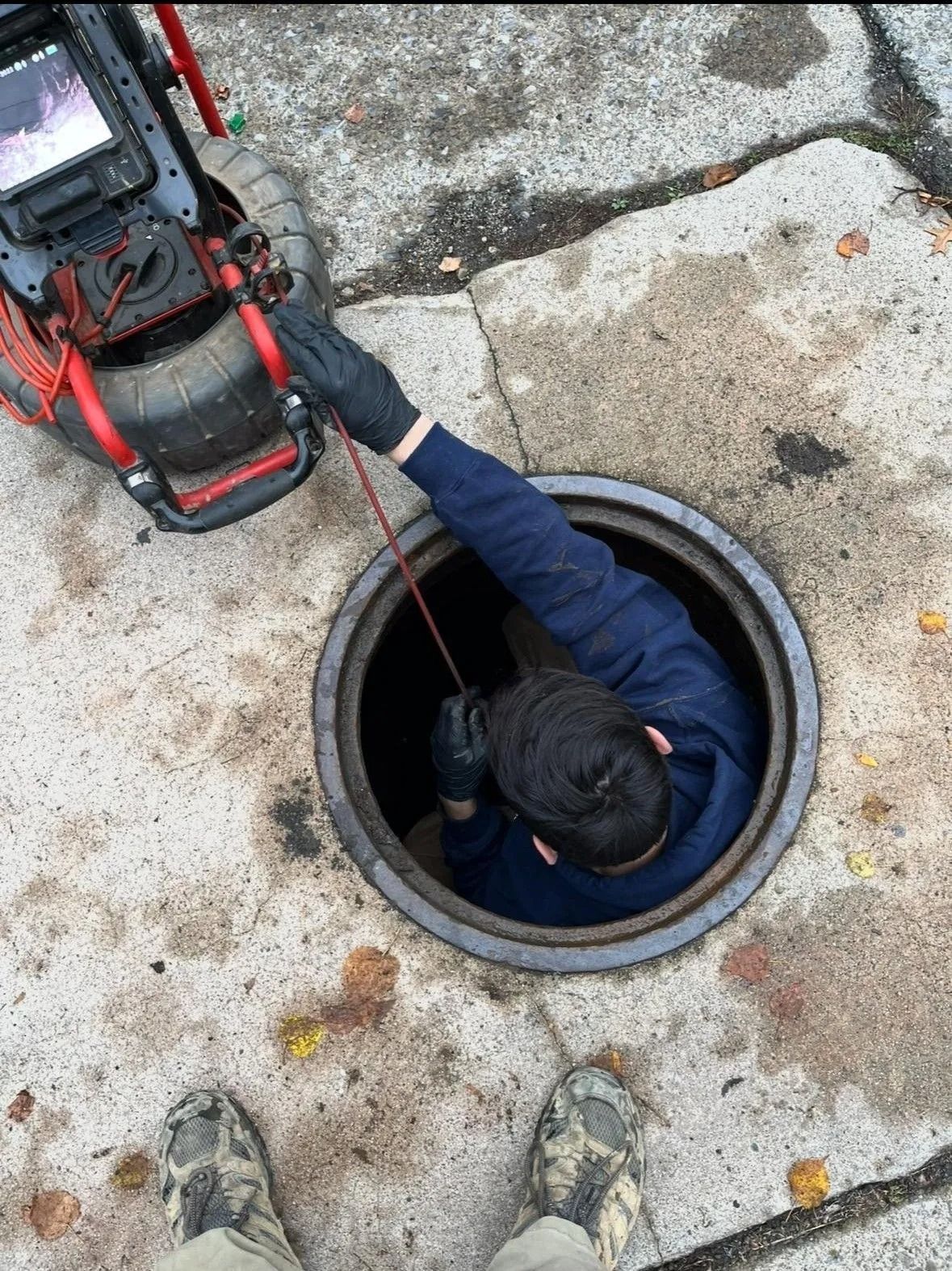Worker climbing from a manhole beside a red-and-black rescue pack, viewed from above