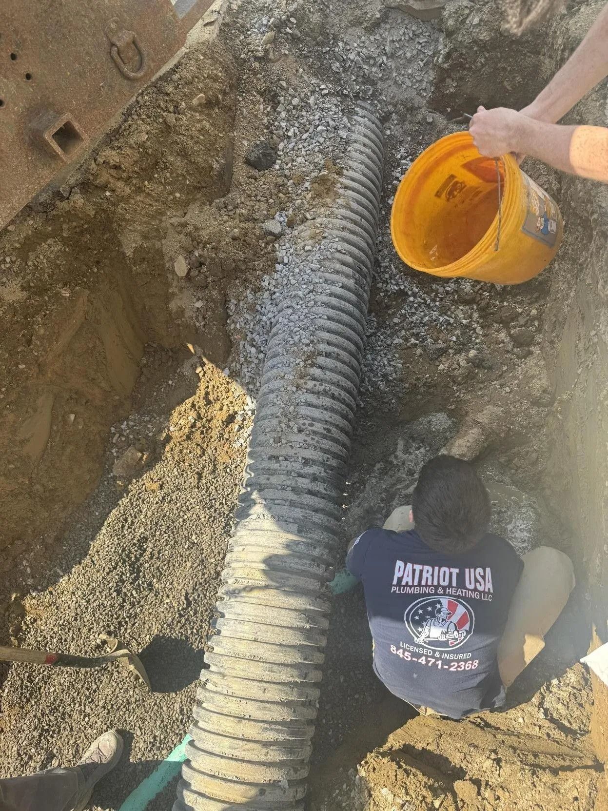 Workers pour orange material into a buried black pipe at a trench site.