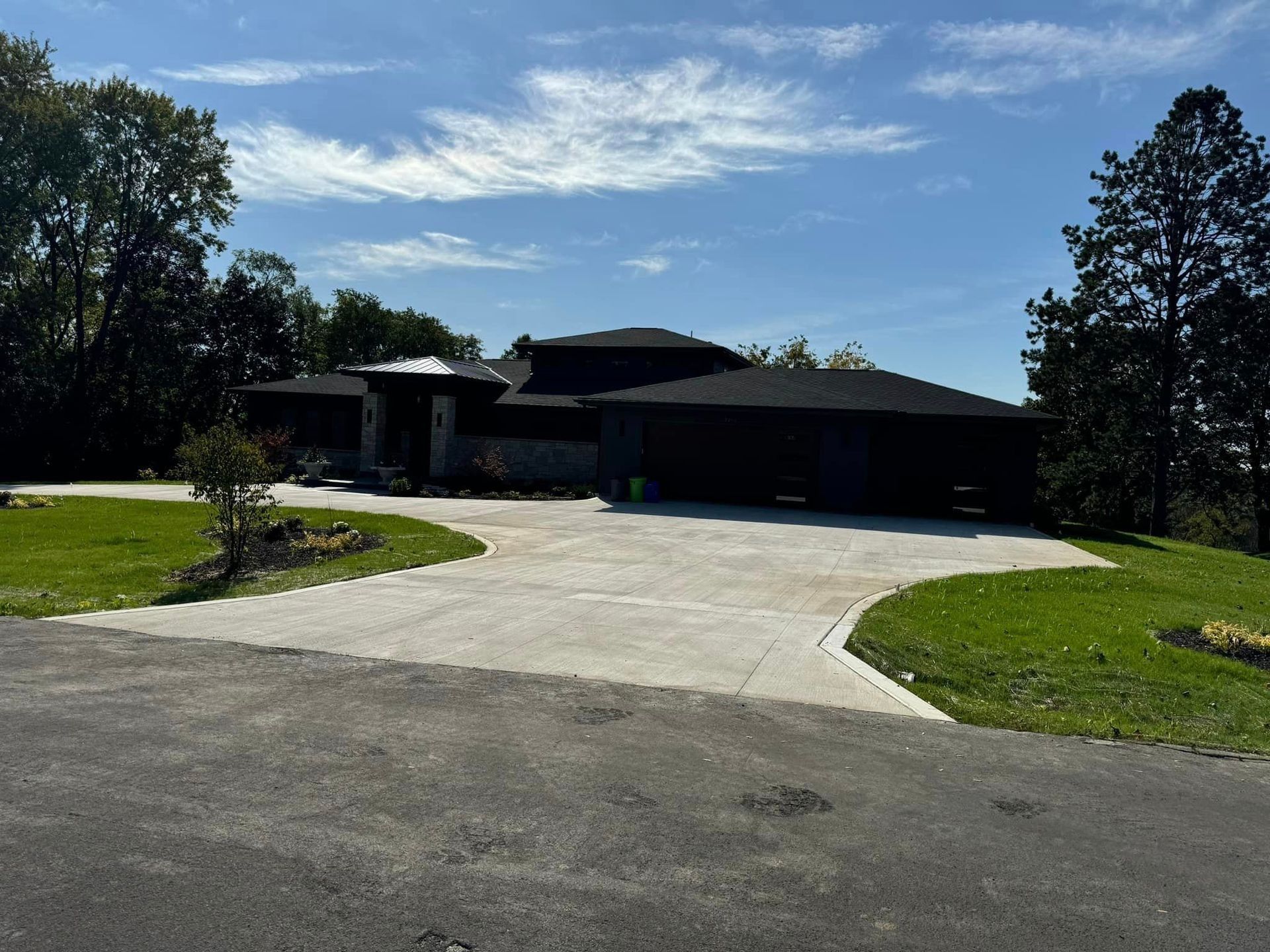 A dark-sided modern house with a large concrete driveway sits on a grassy lawn under a blue sky.