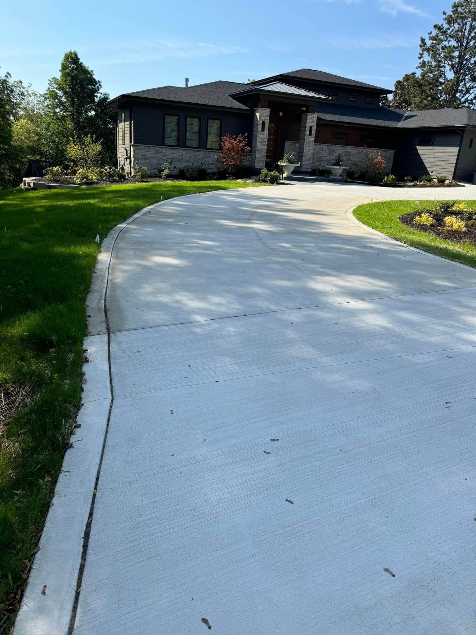 Curved concrete driveway leading to a modern house with a green lawn and trees under a blue sky
