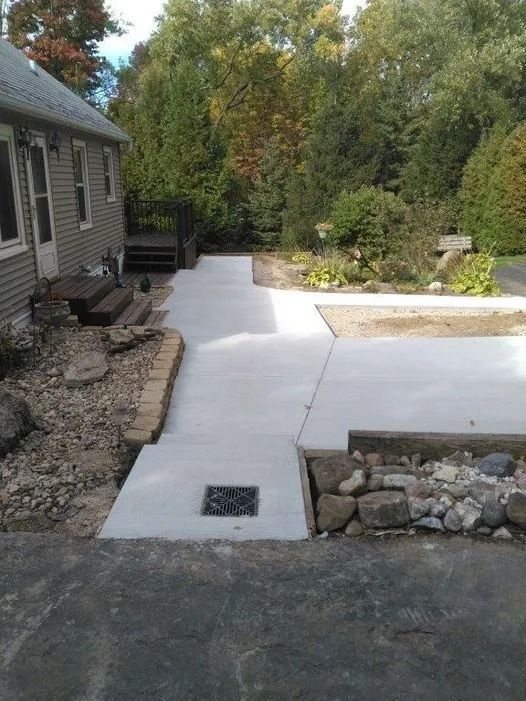 Newly poured concrete sidewalk leading from a house entrance to an outdoor patio with a drain grate in the foreground.