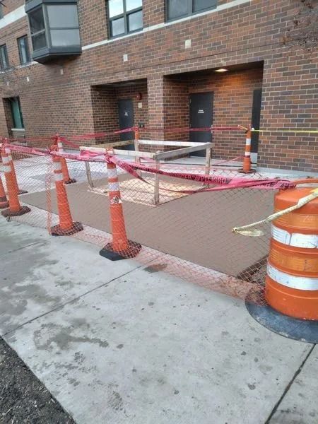 Construction cones and caution tape surround a fenced-off concrete area outside a brick building.