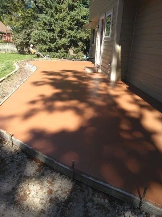 Freshly poured reddish concrete patio beside a house, with sunlight and tree shadows.