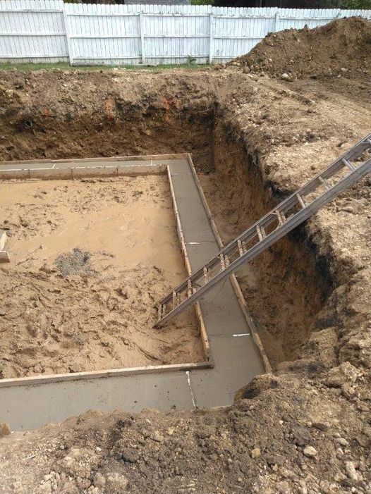 A view into a dirt excavation site showing a partially poured concrete foundation trench with a ladder resting inside.
