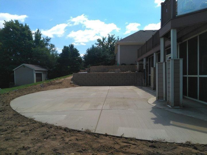 Empty concrete patio beside a building with columns, overlooking a dirt slope and trees under a blue sky