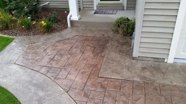 Stamped concrete walkway leading to a front porch and house entrance, bordered by shrubs.