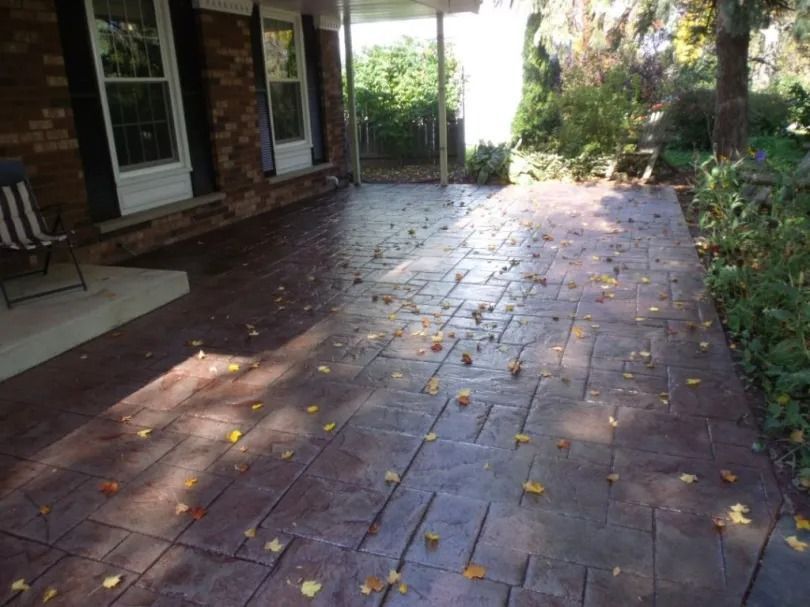 A stamped concrete patio in reddish-brown, with a brick wall, covered porch, and scattered fallen leaves.