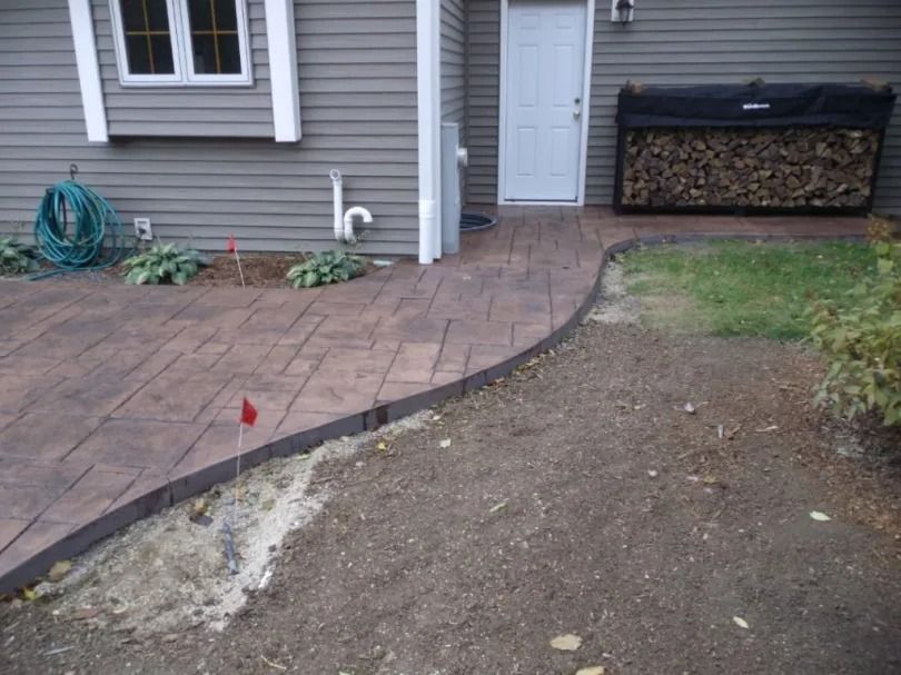 Curved brick patio beside a house, transitioning to bare dirt and grass near a stacked firewood bin.