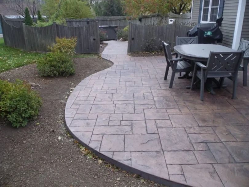 A stamped concrete patio with a dark trim, featuring a table and chairs set near a house next to a wooden fence.