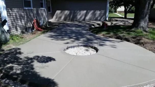 Concrete driveway with a circular fire pit in the center, trees casting shadows nearby.