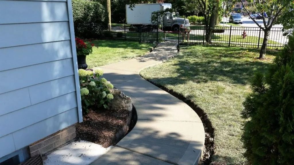 Curved concrete walkway beside blue house, leading through a sunny front yard with trees and a parked truck.