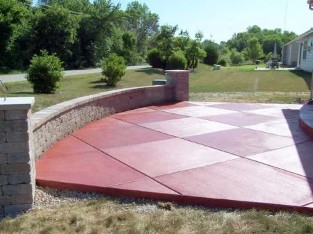 A red, diamond-patterned concrete patio with a curved stone retaining wall in a grassy backyard.