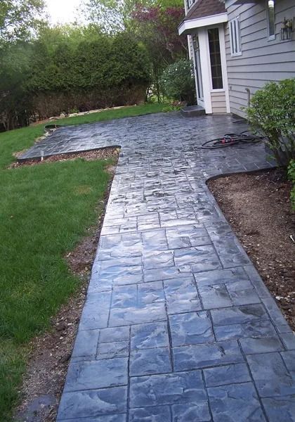 A gray stamped concrete walkway leads to a patio along the side of a house next to a lawn and garden beds.