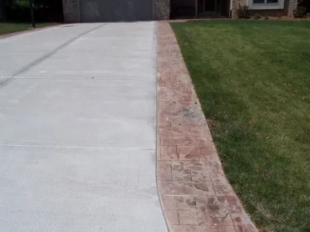 A concrete driveway with a decorative, reddish-brown stamped concrete border running alongside a patch of green grass.