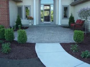 Front entrance walkway with stone patio, white curved path, and potted shrubs by double glass doors
