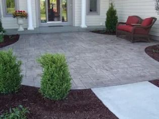 Front walkway of a house with gray paver patio, red chairs, and shrubs in landscaped mulch beds