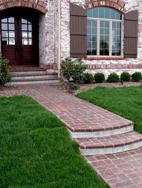 A brick walkway with two steps leads to the front entrance of a house with white brick walls and dark brown shutters.