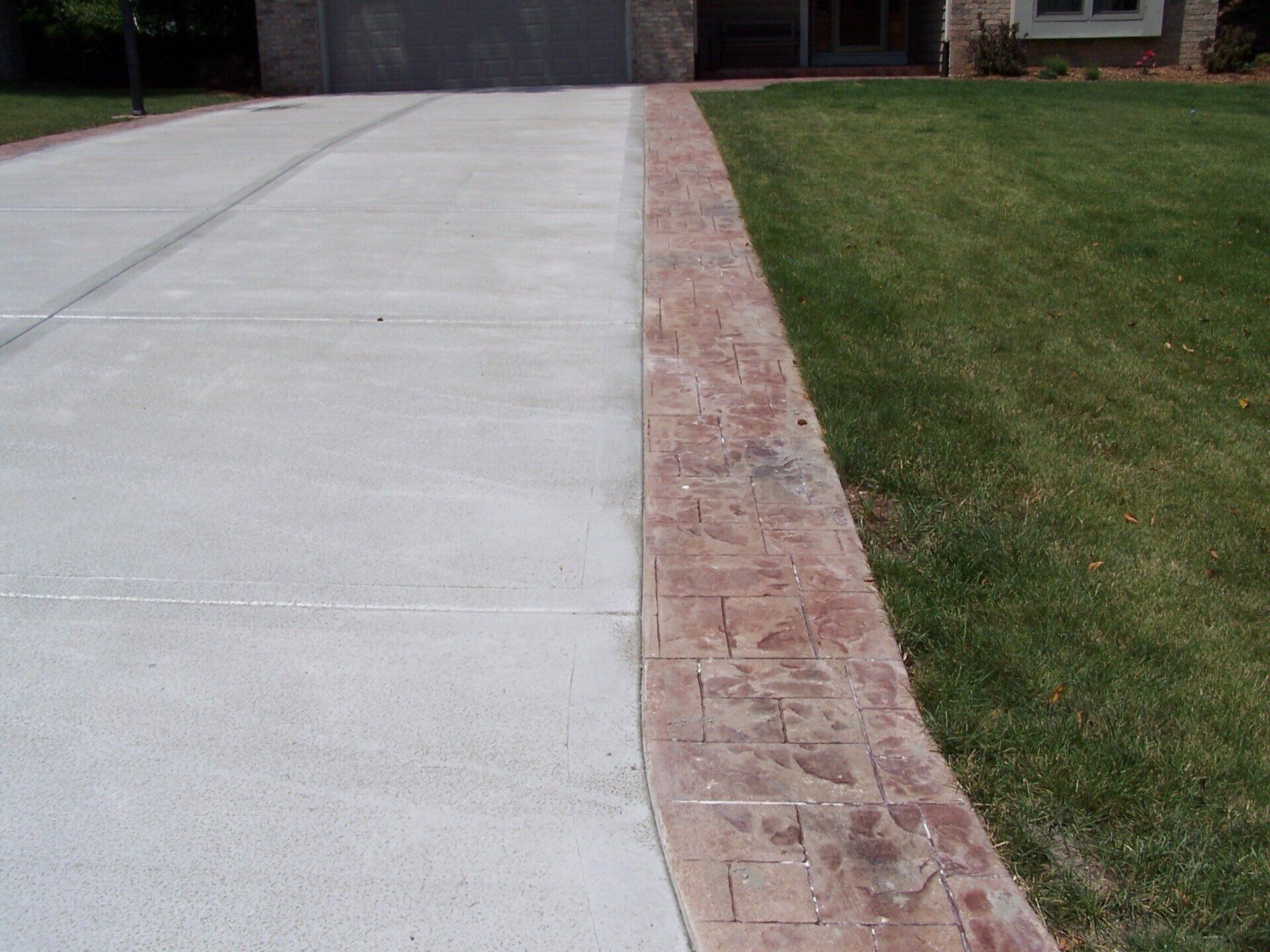 A concrete driveway bordered by a reddish, stamped textured concrete strip next to a green lawn.