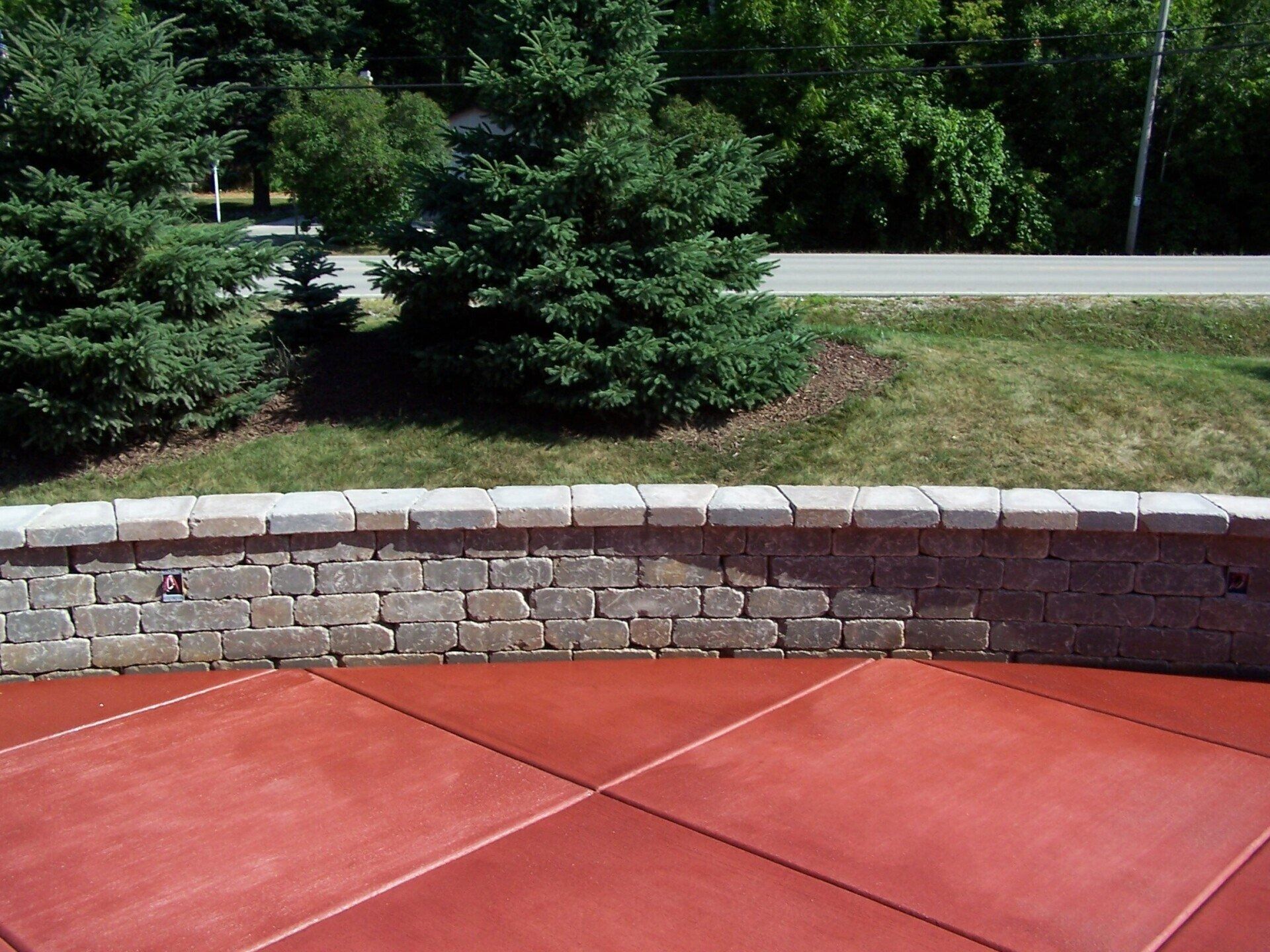 A curved stone retaining wall borders a red concrete patio, with green pine trees and a road in the background.