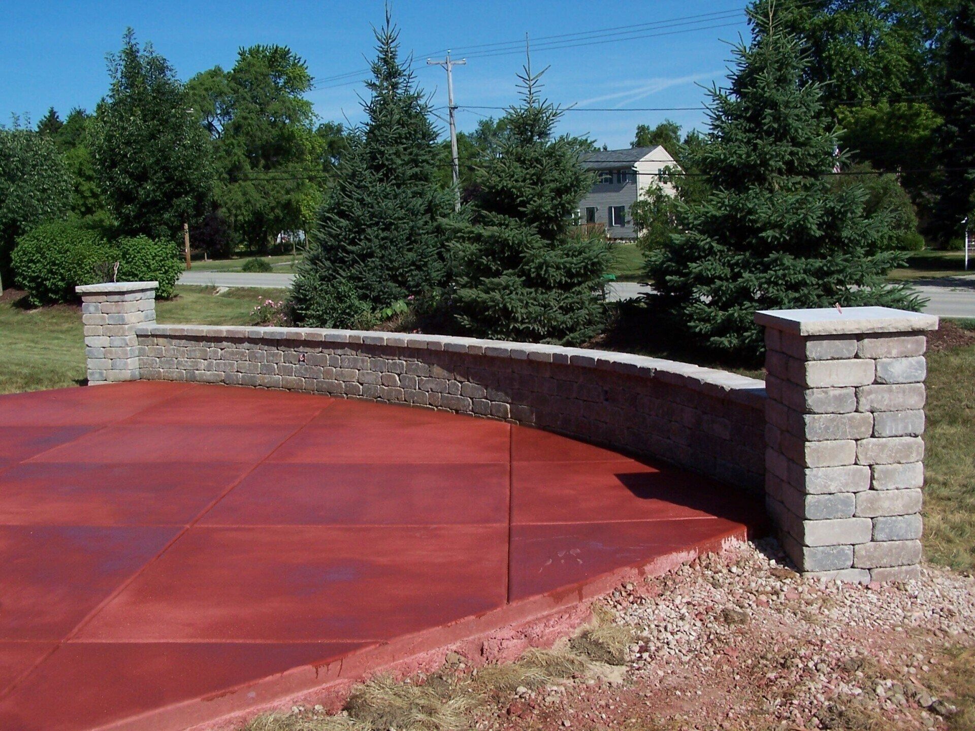 A red-tiled patio with a curved stone retaining wall flanked by two brick pillars in a grassy landscape with pine trees.
