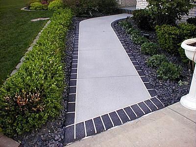 A concrete walkway bordered with dark bricks, flanked by green hedges and grey stone landscaping.