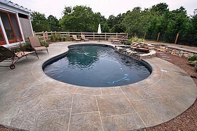 A kidney-shaped swimming pool with a stone patio, loungers, a rock waterfall, and a wooden fence in a backyard setting.