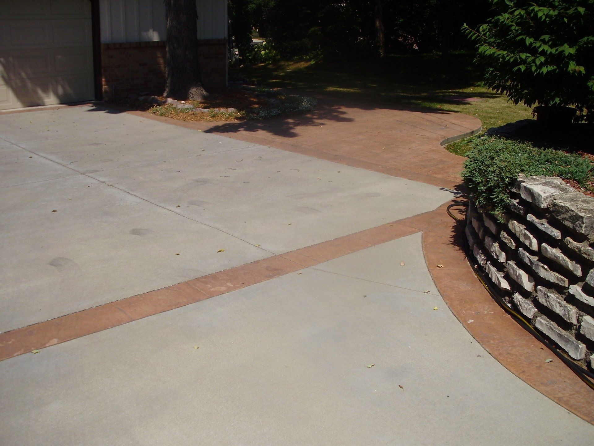 A driveway with gray concrete sections bordered by reddish-brown decorative paving near a stone retaining wall.