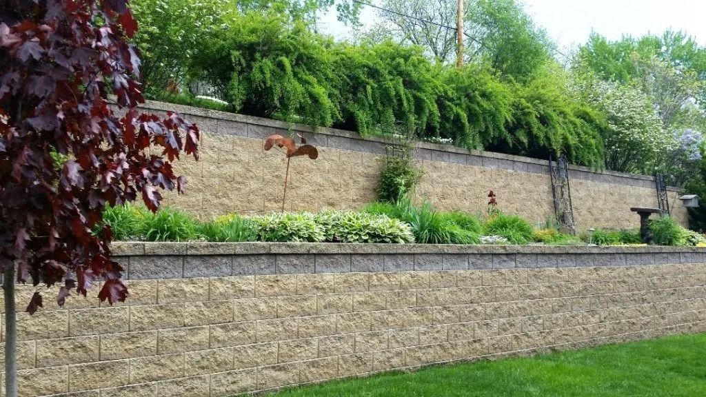 A two-tiered stone retaining wall with lush green landscaping, shrubs, and a dark-leaved tree in the foreground.