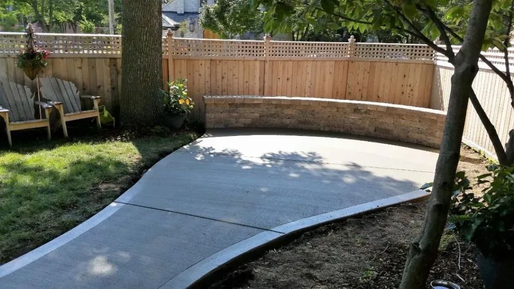 Curved concrete patio beside a wooden fence and trees in a backyard.