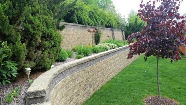 A landscaped yard with a curved stone retaining wall, lush green lawn, trees, shrubs, and a tall stone fence in the back.